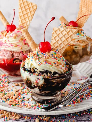 Group of three sundaes, classic, strawberry and caramel, with whipped cream, nuts, sprinkles, wafers and cherry, on a plate with spoons, nuts and sprinkles.
