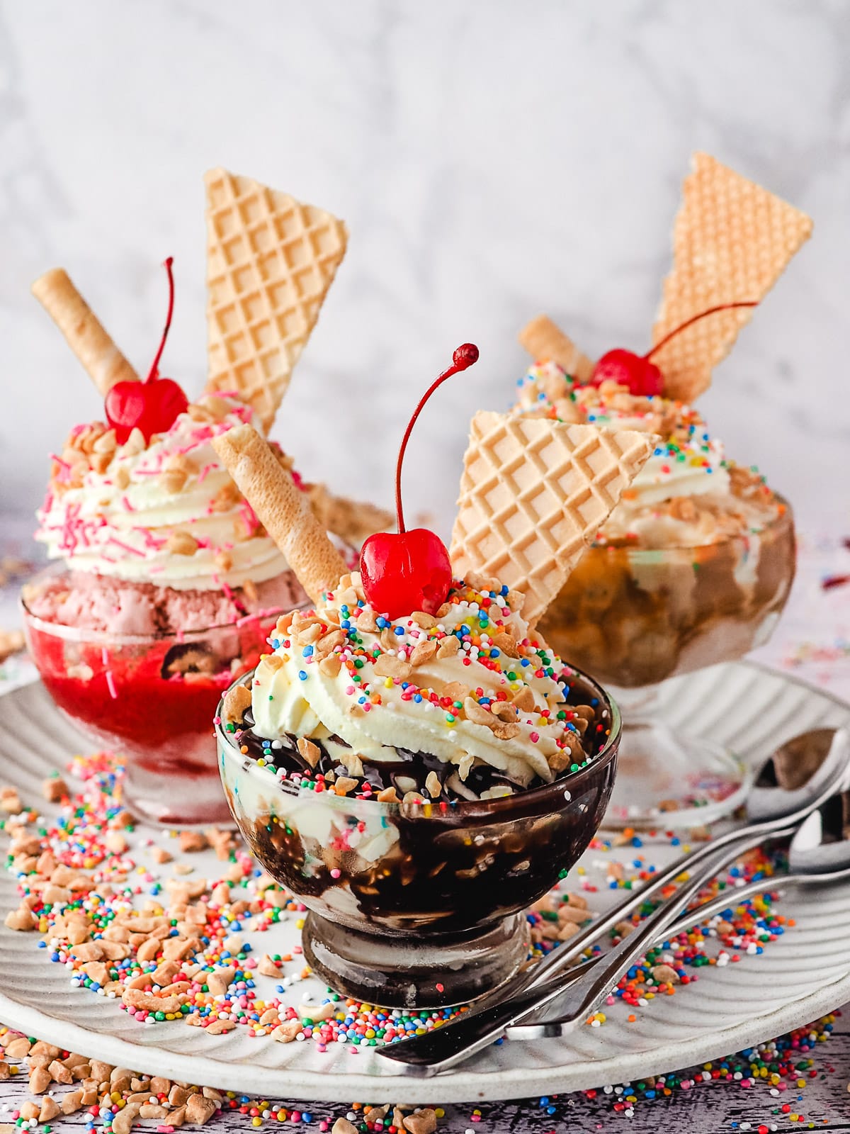 Group of three sundaes, classic, strawberry and caramel, with whipped cream, nuts, sprinkles, wafers and cherry, on a plate with spoons, nuts and sprinkles.
