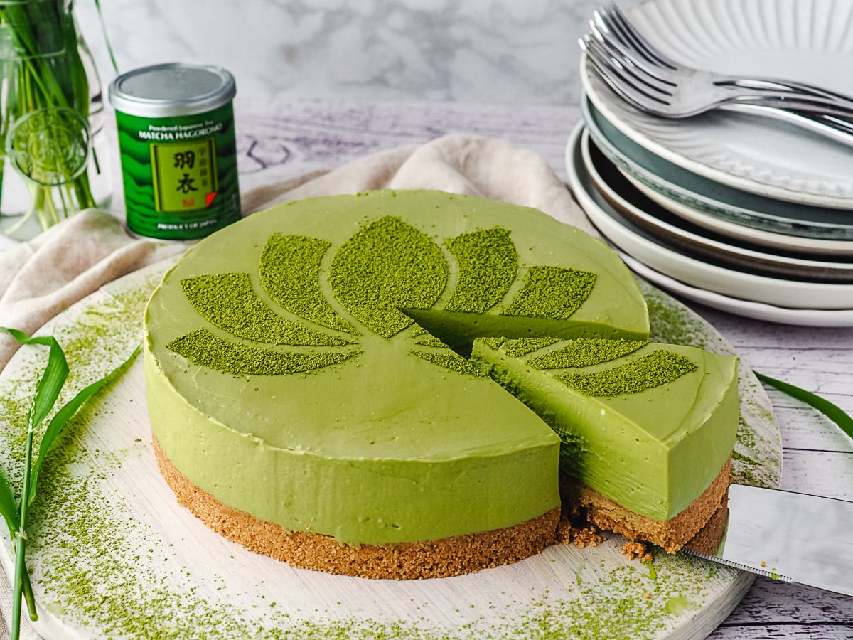 Slicing cheesecake, with jar of green tea and stack of plates in background.