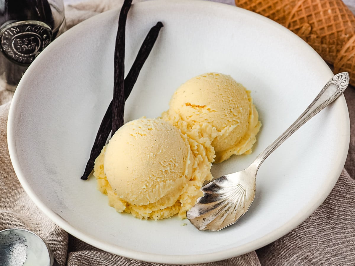 Two scoops of ice cream in a bowl, with vanilla beans and vintage spoon on the side, and ice cream cones and scoop in background.