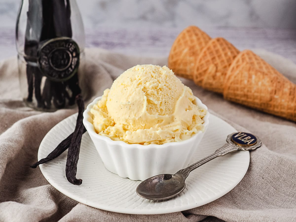 Single scoop of ice cream in a cone in a vintage jar, with vanilla beans and ice cream cones in background.