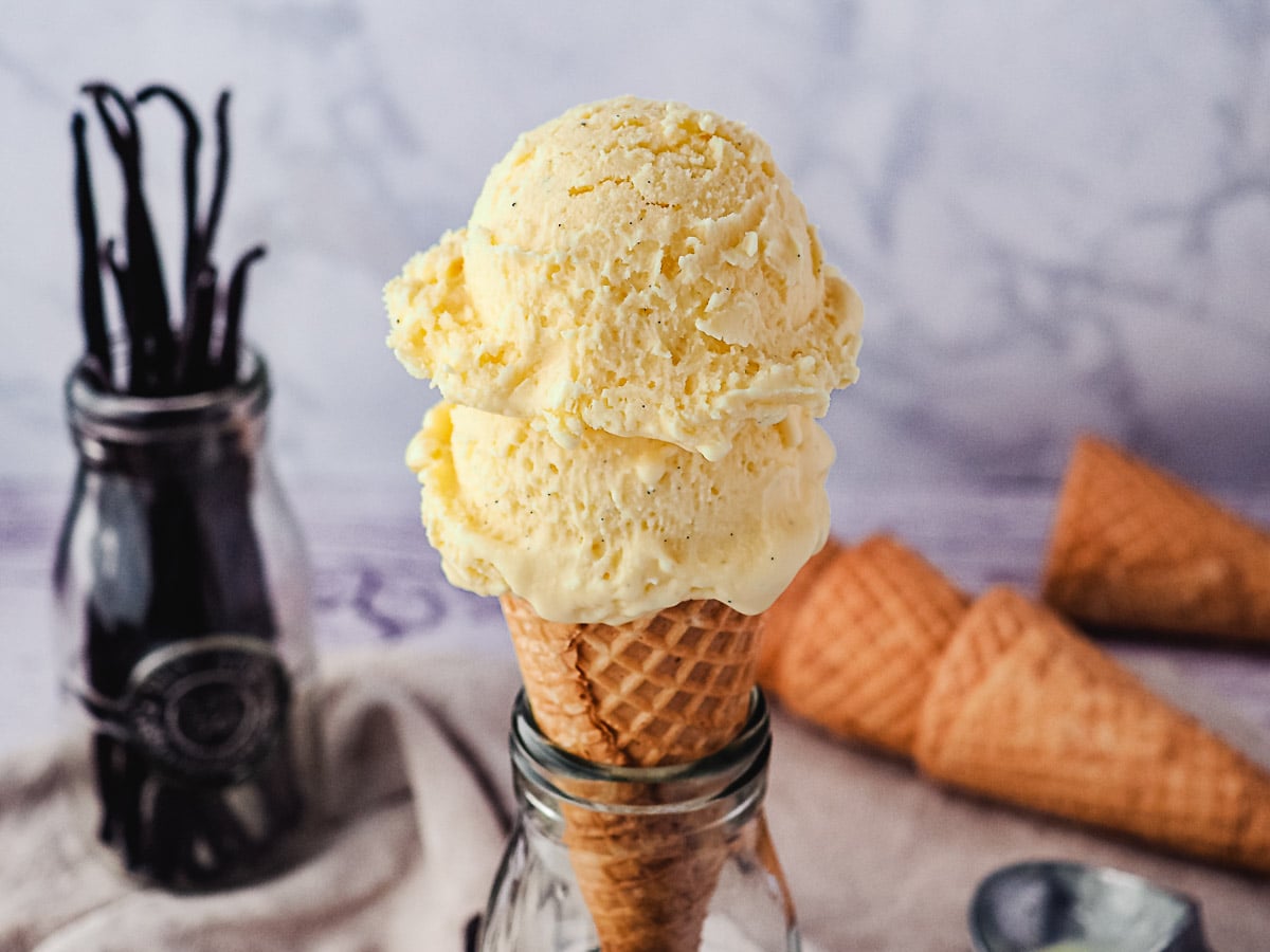 Two scoops of ice cream in a cone in a vintage jar, with vanilla beans and ice cream cones in background.