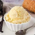 Scoop of ice cream in a bowl, with vanilla beans and spoon on the side and ice cream cones in background.