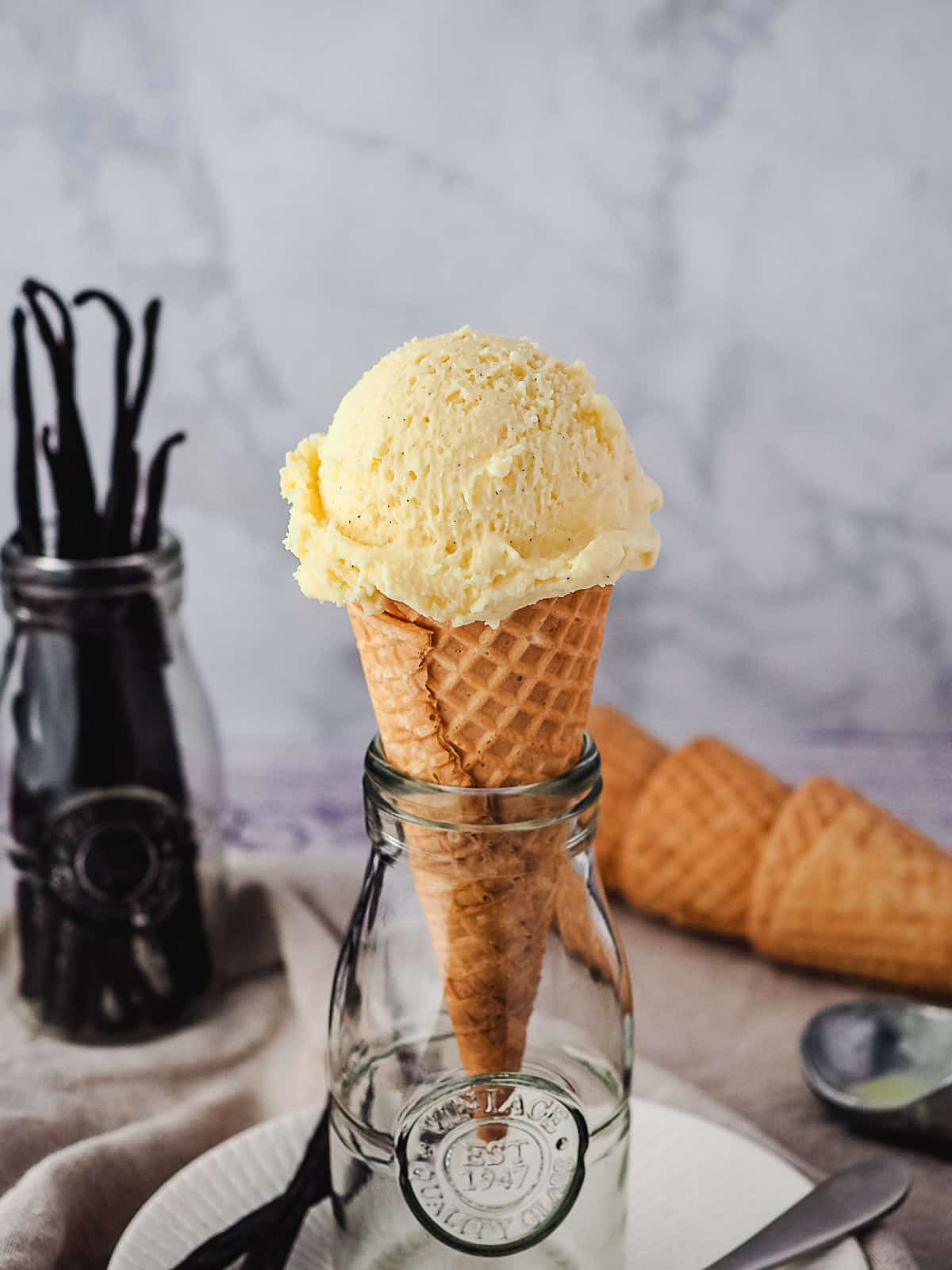 Single scoop of ice cream in a cone in a vintage jar, with vanilla beans and ice cream cones in background.