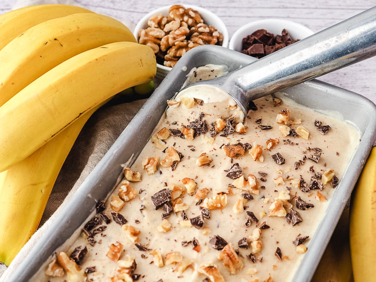 Scooping ice cream from a pan, with bananas, walnuts and chocolate chunks on the side.