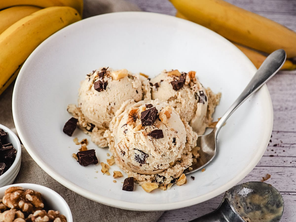 Three scoops of ice cream in a bowl with a spoon and fresh bananas, walnuts and chocolate chunks on the side.