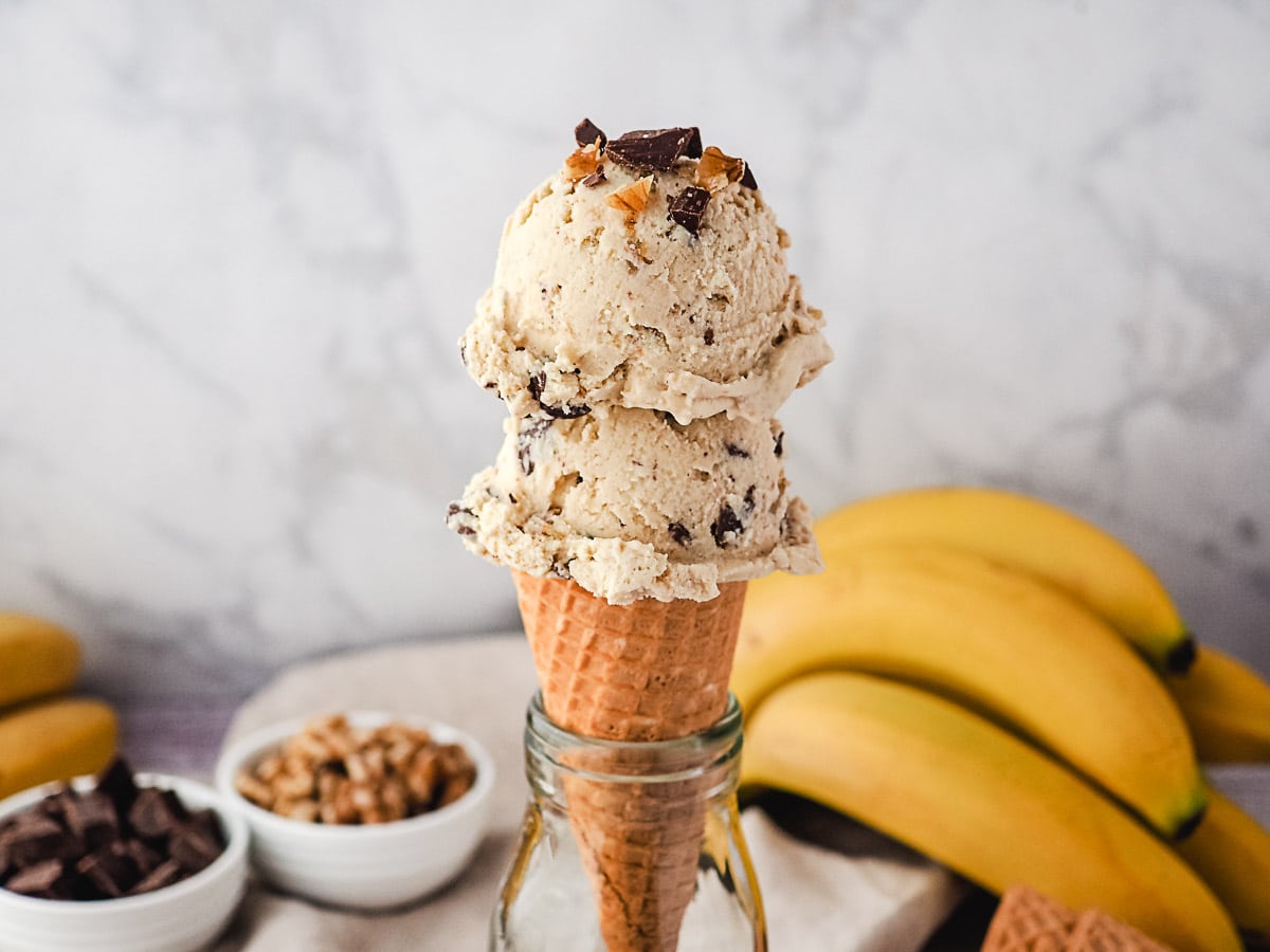 Two scoops of ice cream in a cone, with bananas, walnuts and chocolate chunks in the background.