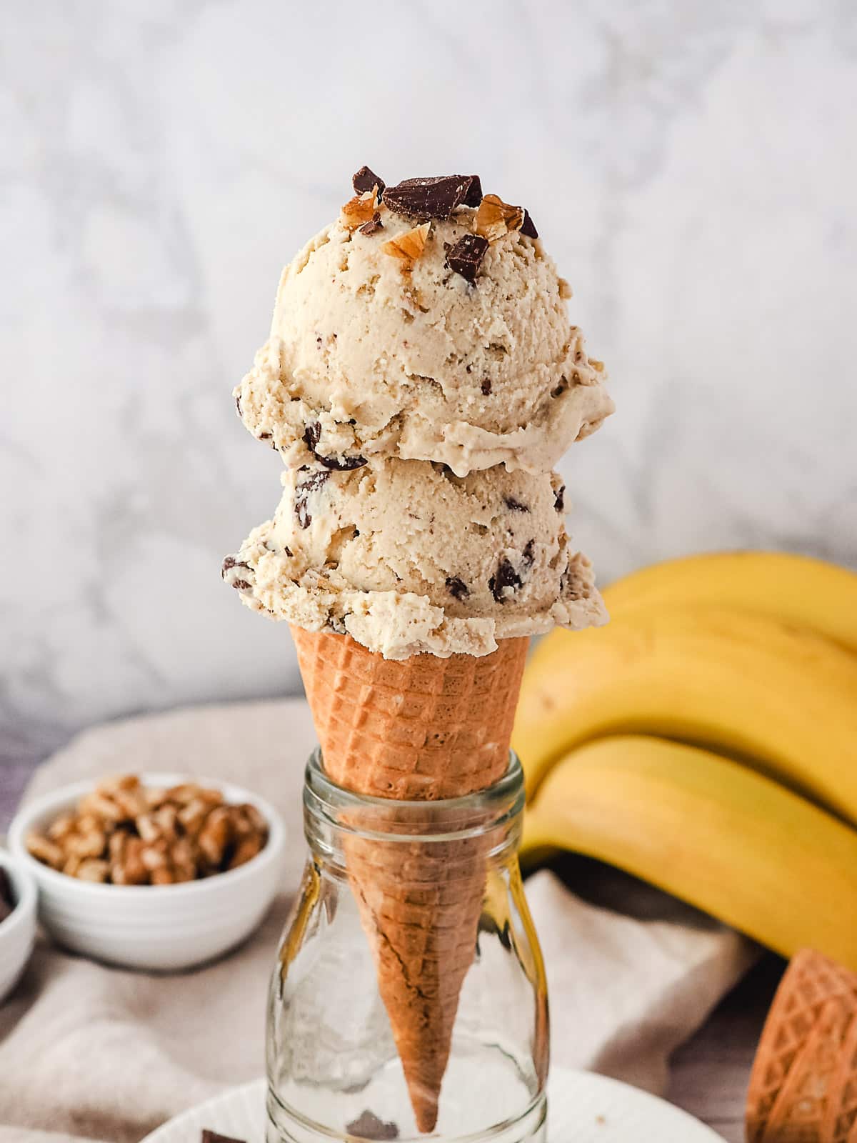 Two scoops of ice cream in a cone, with bananas, walnuts and chocolate chunks in the background.