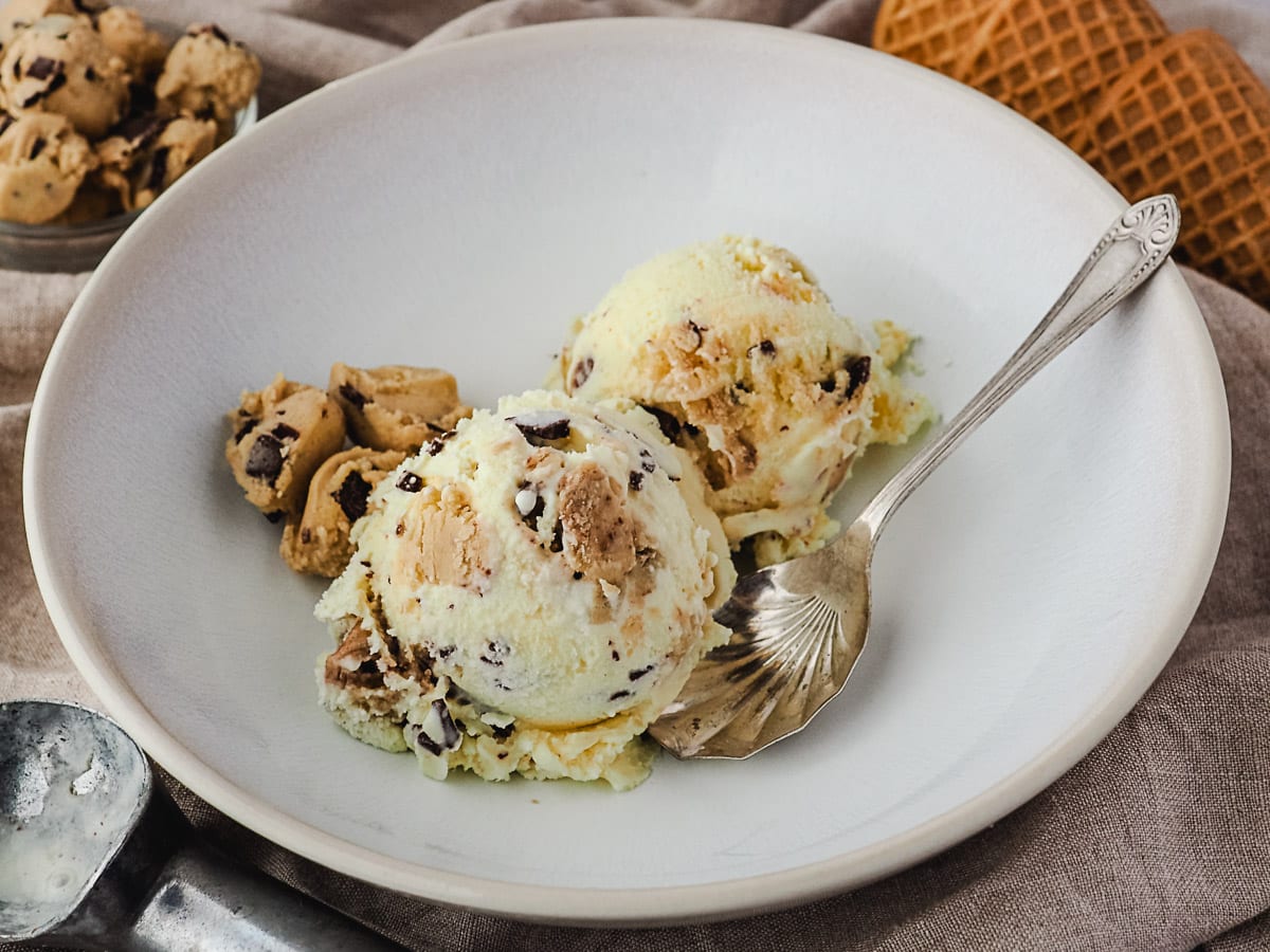 Two scoops of ice cream in a bowl with ice cream scoop, bowl of cookie dough pieces and ice cream cones in the background.