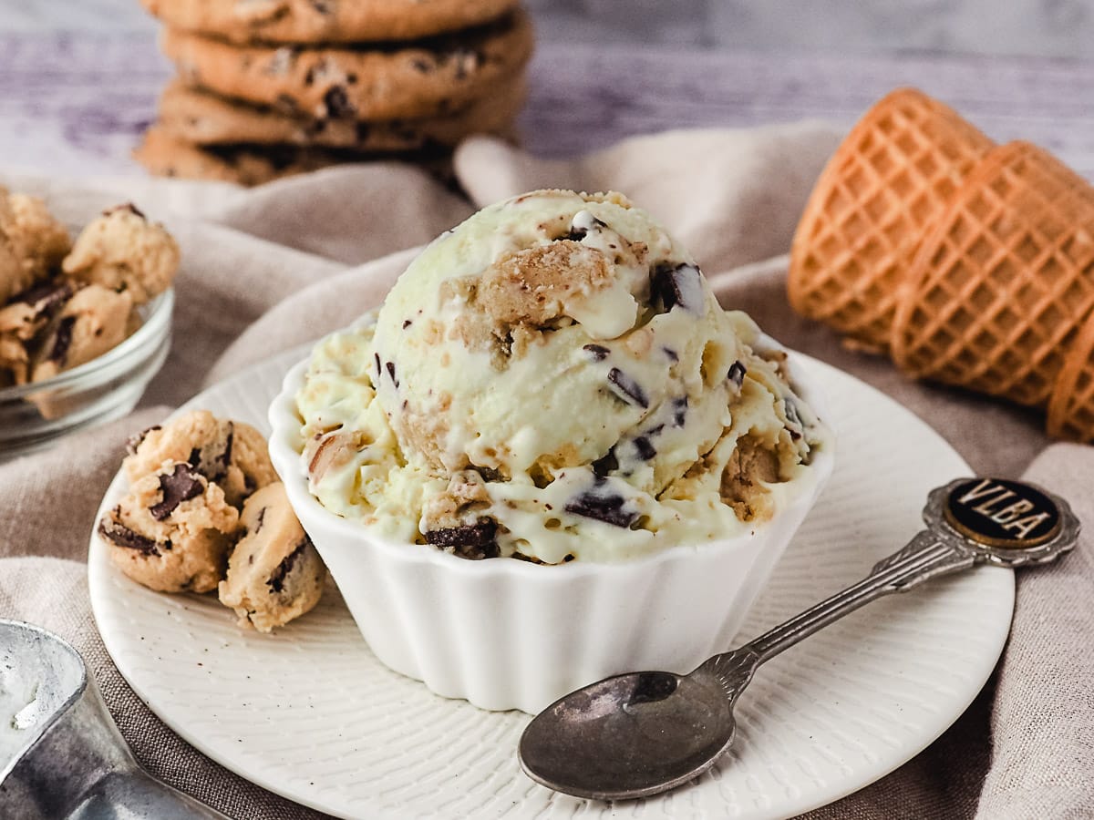 Scoop of ice cream in a bowl with a spoon and pieces of edible cookie dough on the side and ice cream cones in the background.