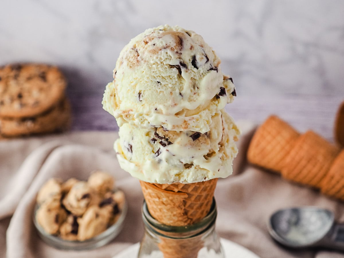 Two scoops of ice cream in a cone, with edible cookie dough and ice cream cones in background.
