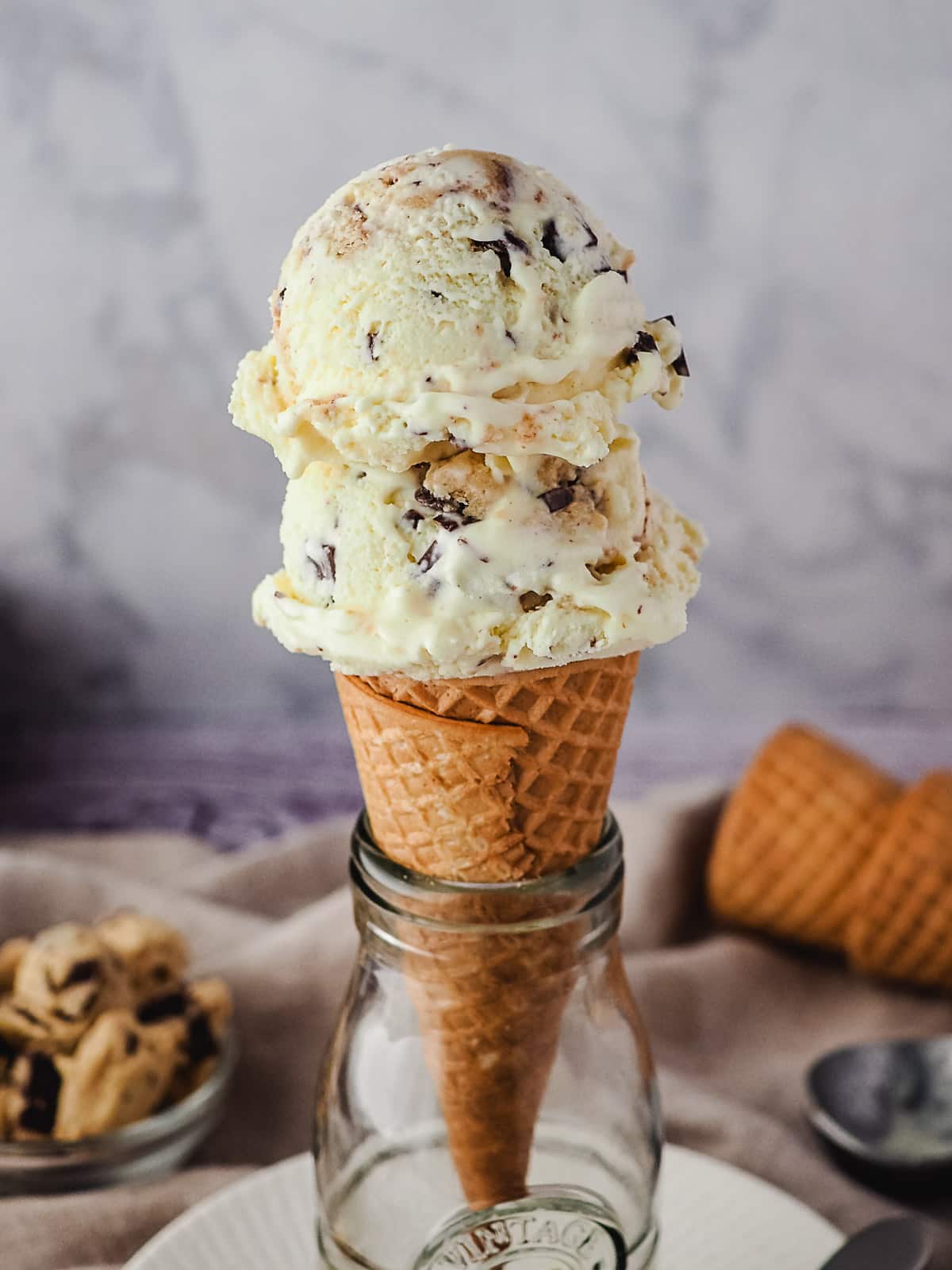 Two scoops of ice cream in a cone, with edible cookie dough and ice cream cones in background.