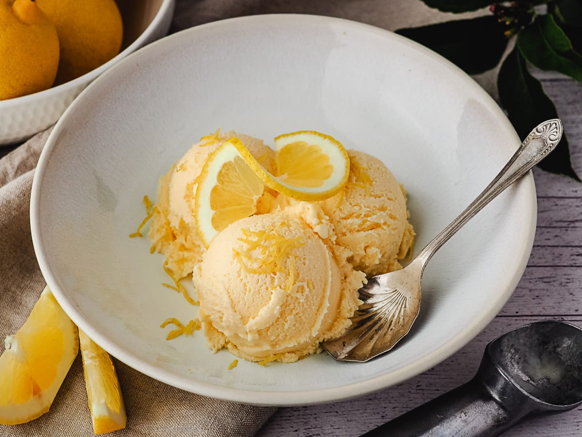 Three scoops of ice cream in a bowl with a spoon on the side and fresh lemons and ice cream scoop in the background.