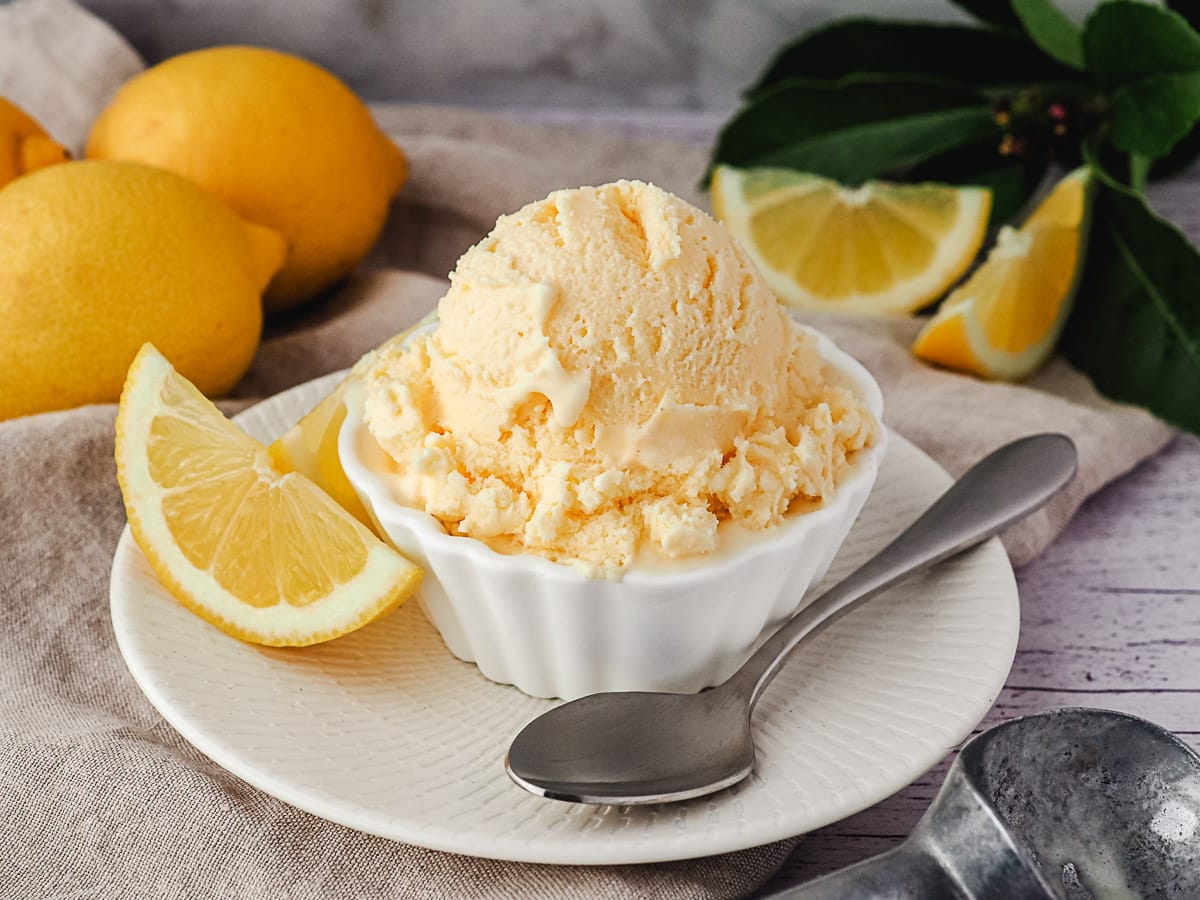 Scoop of ice cream in a bowl with spoon on the side, with fresh lemons and ice cream scoop in the background.