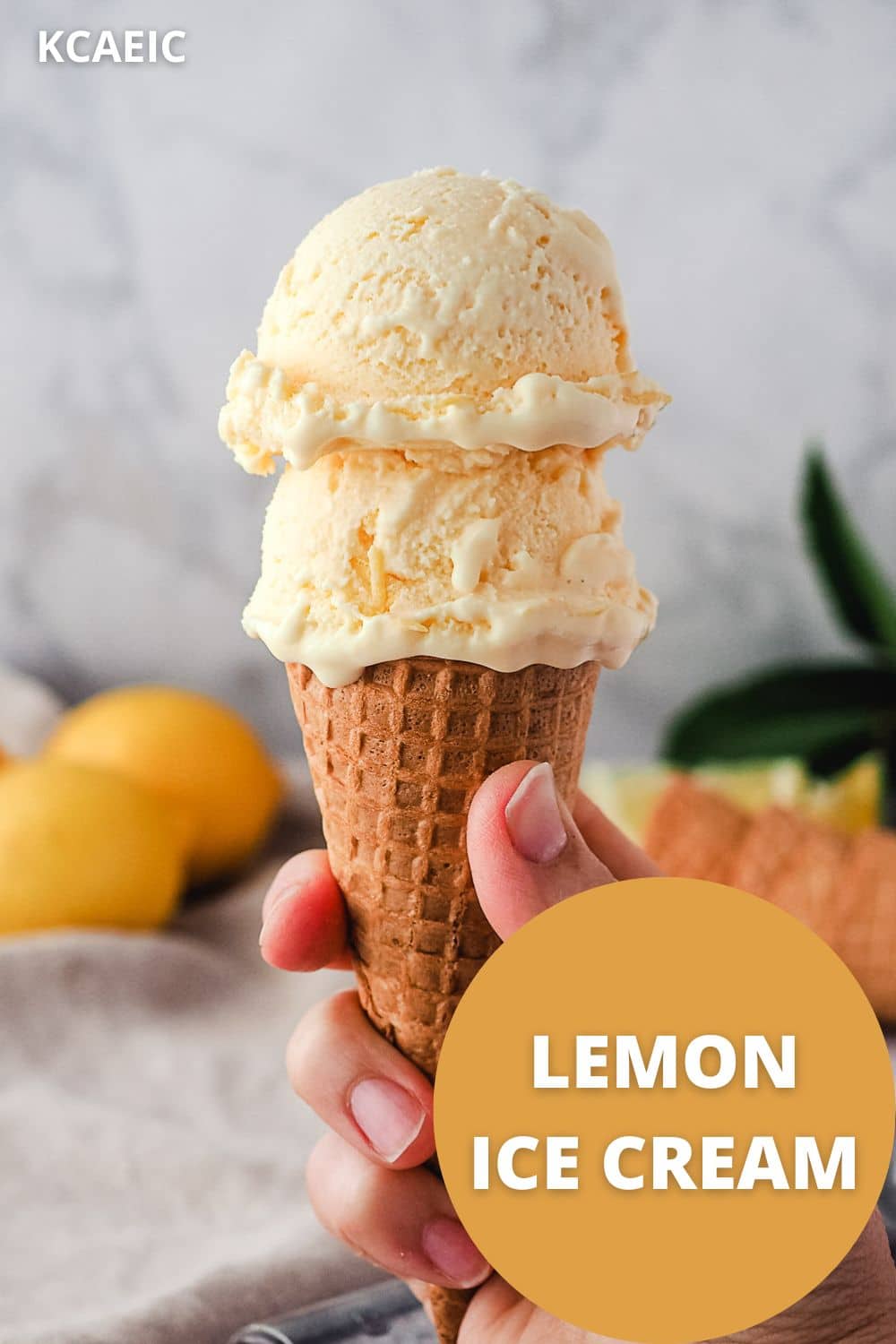 Hand holding two scoops of ice cream in a cone with lemons in the background and text overlay, lemon ice cream.