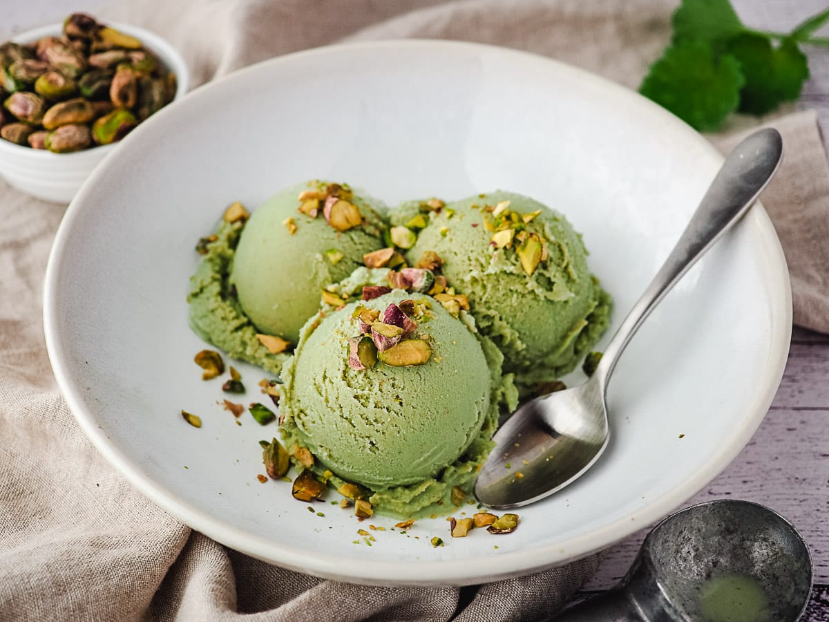 Three scoops of ice cream in a bowl topped with chopped roasted pistachios, with a spoon on the side.