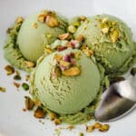 Three scoops of ice cream in a bowl topped with chopped roasted pistachios, with a spoon on the side.