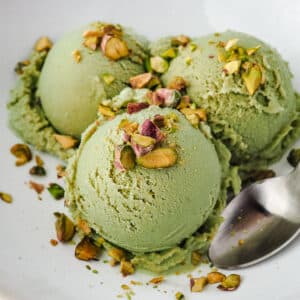Pistachio Ice Cream 9 Three scoops of ice cream in a bowl topped with chopped roasted pistachios, with a spoon on the side.