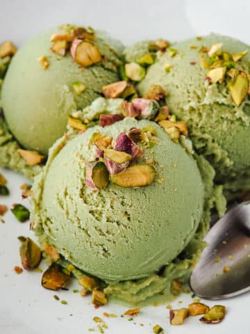 Three scoops of ice cream in a bowl topped with chopped roasted pistachios, with a spoon on the side.