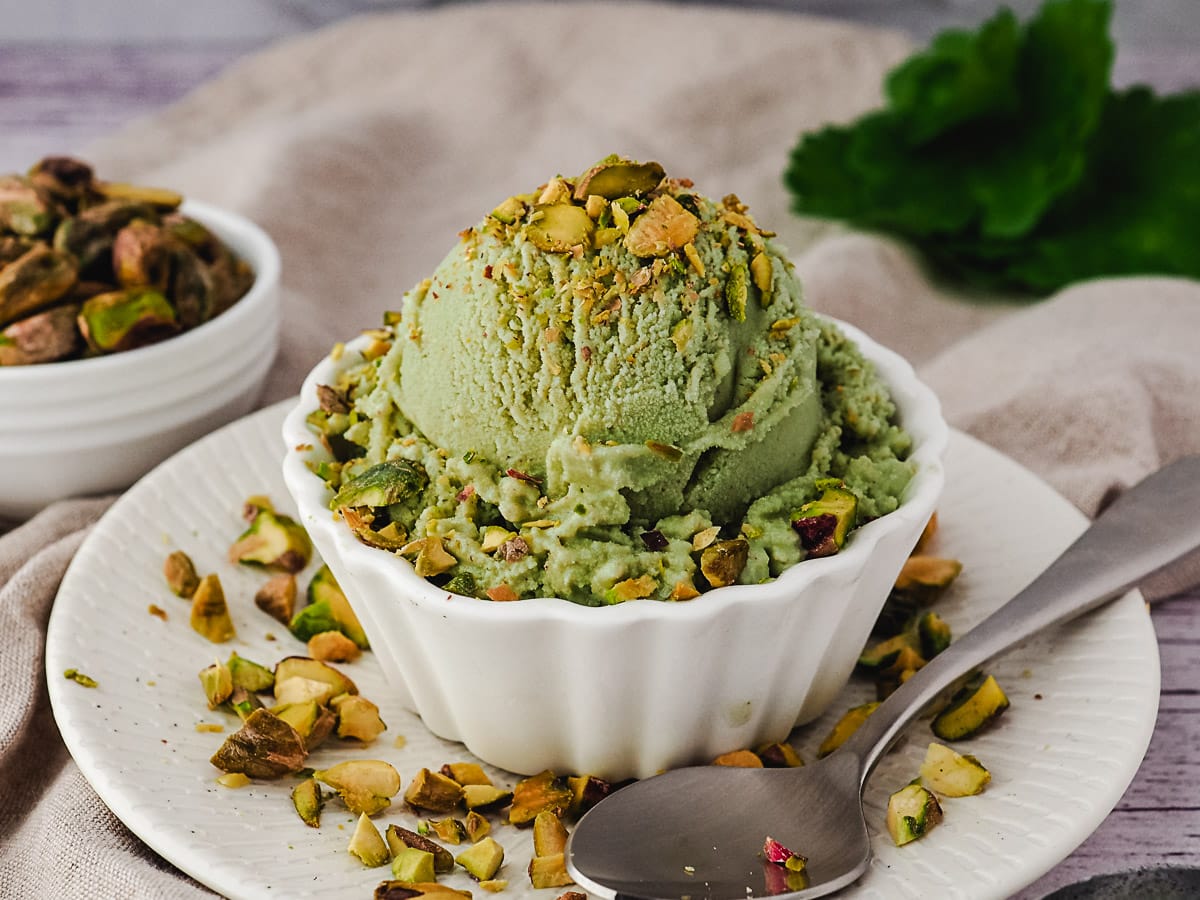 Scoop of ice cream in a bowl topped with pistachio pieces, with a spoon and extra pistachios on the side.