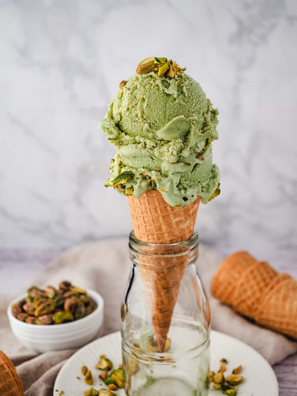 Two scoops of ice cream topped with pistachio pieces in a cone balanced in a glass jar, with pistachios and ice cream cones in the background.
