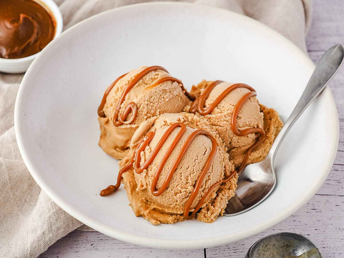 Three scoops of ice cream in a bowl with dulce de leche drizzle and dulce de leche in background.