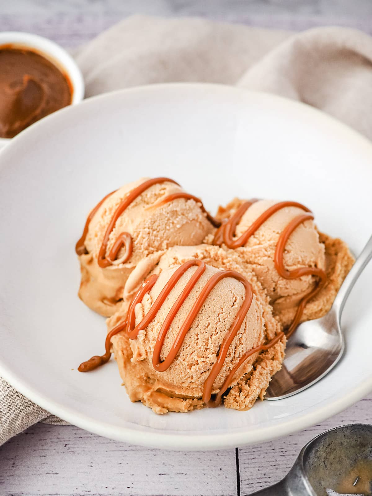 Three scoops of ice cream in a bowl with dulce de leche drizzle and dulce de leche in background.