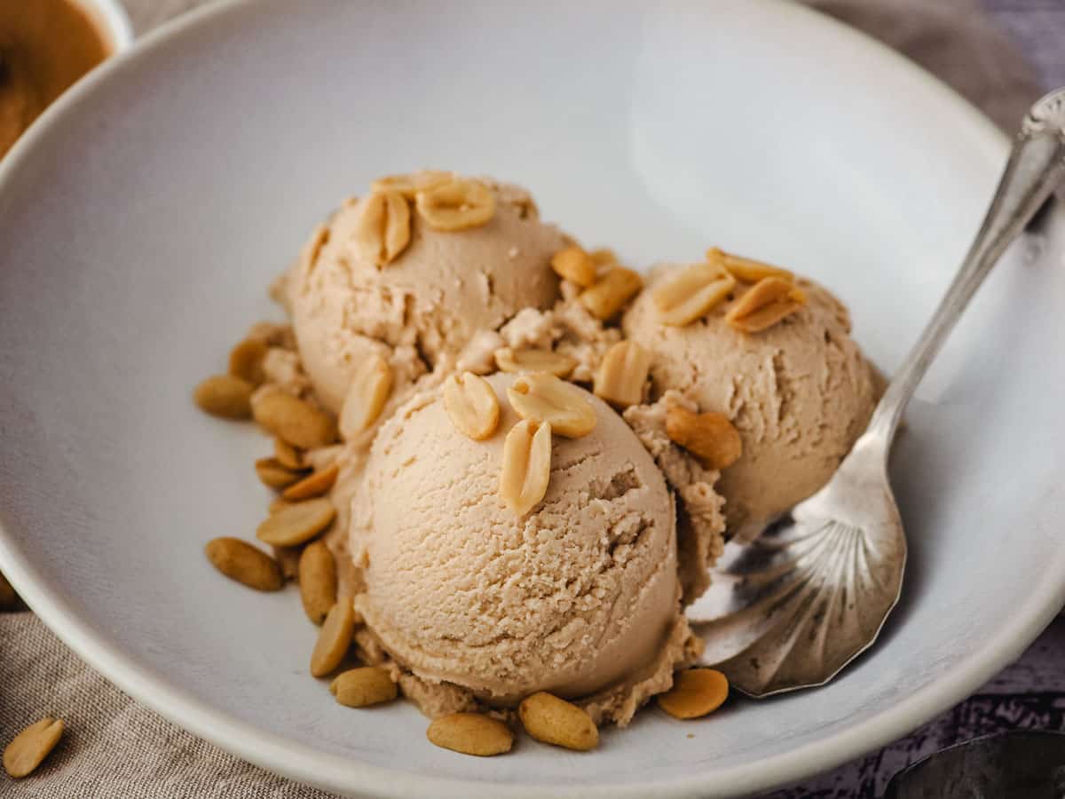 Peanut butter ice cream in a bowl with roasted peanuts and a spoon on the side.