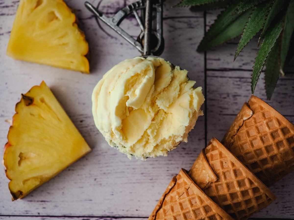 Pineapple Ice Cream 7 Ice cream in an ice cream scoop, with fresh pineapple and ice cream cones on the side.