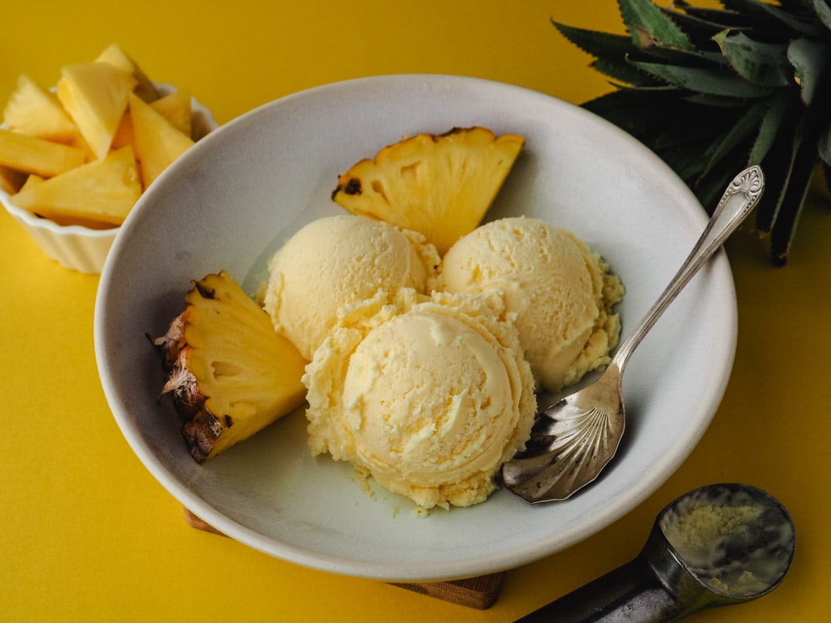 Three scoops of ice cream in a bowl with a spoon and fresh pineapple on the side.