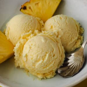 Close up three scoops of ice cream in a bowl with a spoon and fresh pineapple on the side.