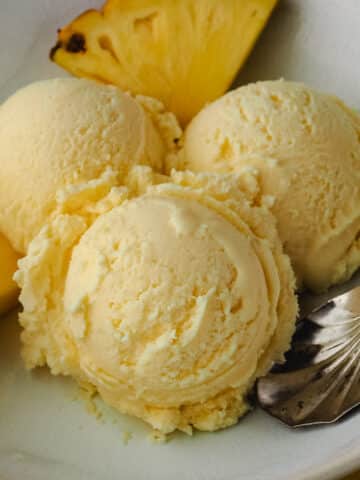 Close up three scoops of ice cream in a bowl with a spoon and fresh pineapple on the side.