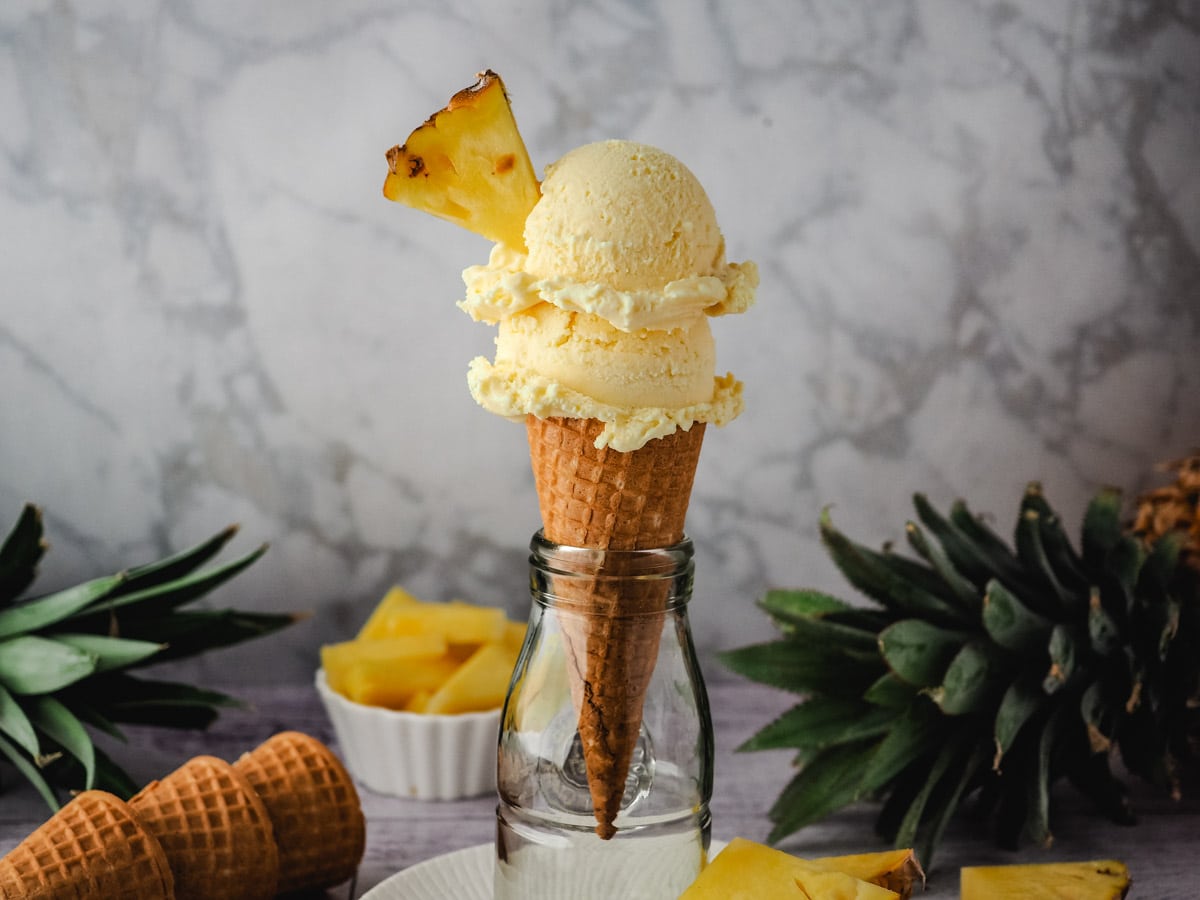 Pineapple Ice Cream 2 Two scoops of ice cream in a cone garnished with fresh pineapple, with fresh pineapple in the background.