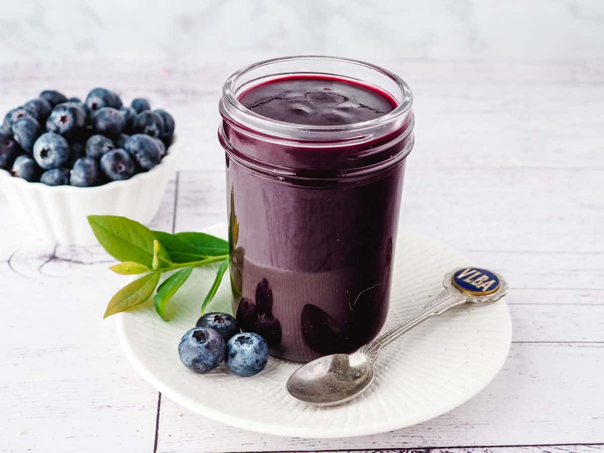 Open mason jar of blueberry coulis on a plate with a spoon on the side, and fresh blueberries in the background.