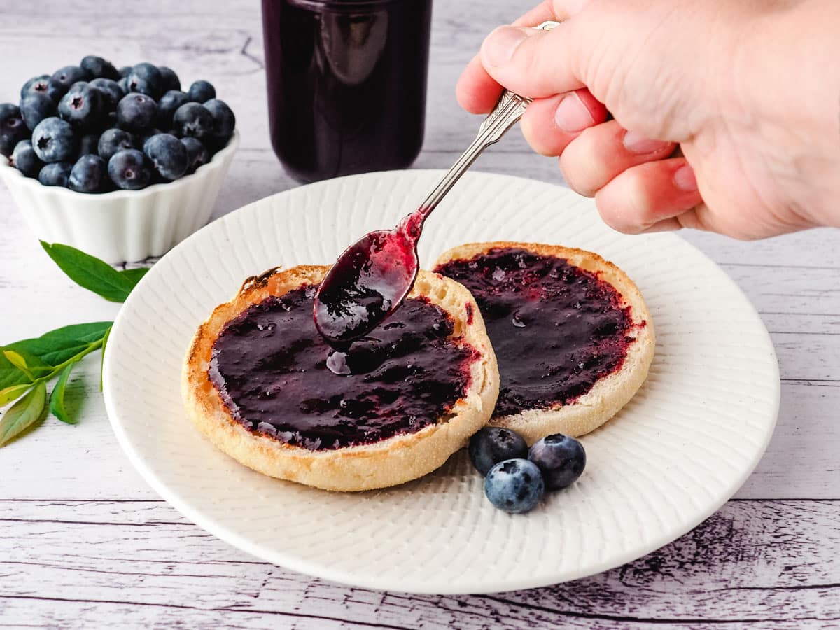 Spooning blueberry coulis onto toasted English muffins, with jar of coulis and fresh blueberries in the background.