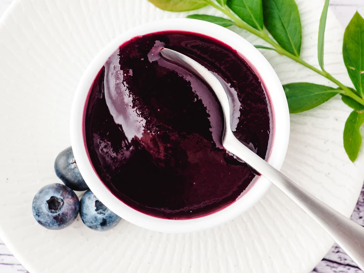 Blueberry Coulis 2 Blueberry coulis in a serving bowl with a spoon, and fresh blueberries and blueberry leaves on the side.