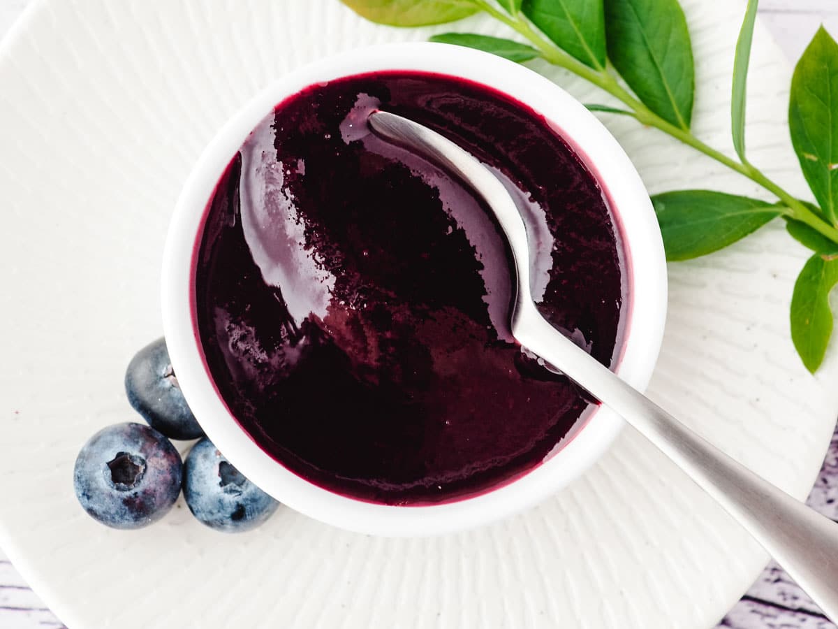 Blueberry coulis in a serving bowl with a spoon, and fresh blueberries and blueberry leaves on the side.