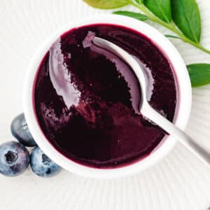 Blueberry coulis in a serving bowl with a spoon, and fresh blueberries and blueberry leaves on the side.