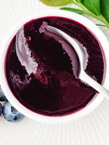 Blueberry coulis in a serving bowl with a spoon, and fresh blueberries and blueberry leaves on the side.