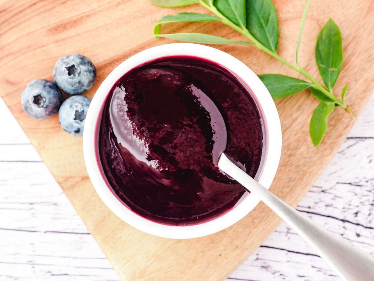 Blueberry coulis in a serving bowl with a spoon, and fresh blueberries and blueberry leaves on the side.