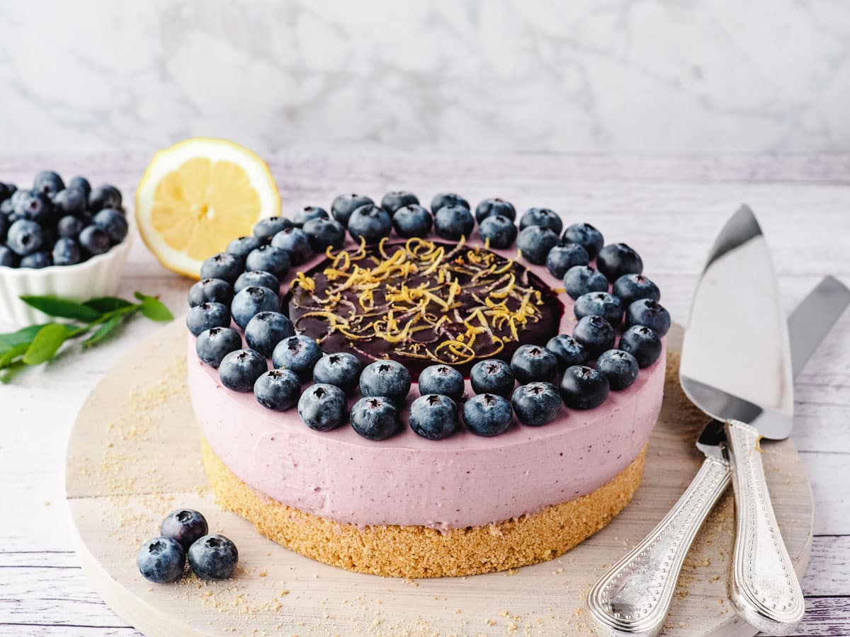 Whole no bake blueberry cheesecake decorated with fresh blueberries, blueberry coulis and lemon zest, on a boar with serving wear and fresh blueberries on the side.