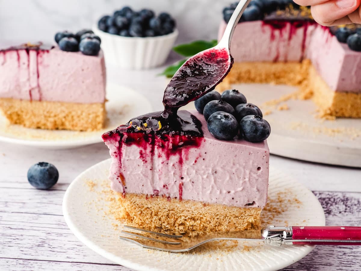 Blueberry coulis being spooned onto a slice of no bake blueberry cheesecake.