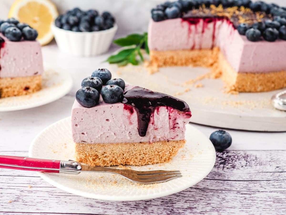 Slice of blueberry cheesecake with blueberry coulis, with another slice of cake, rest of cake and fresh blueberries in the background.