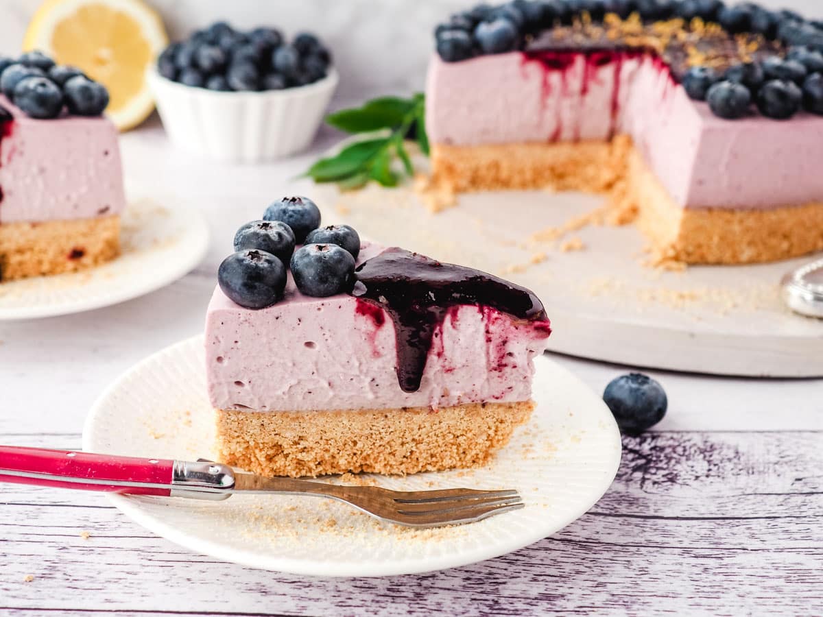 No Bake Blueberry Cheesecake 10 Slice of blueberry cheesecake with blueberry coulis, with another slice of cake, rest of cake and fresh blueberries in the background.