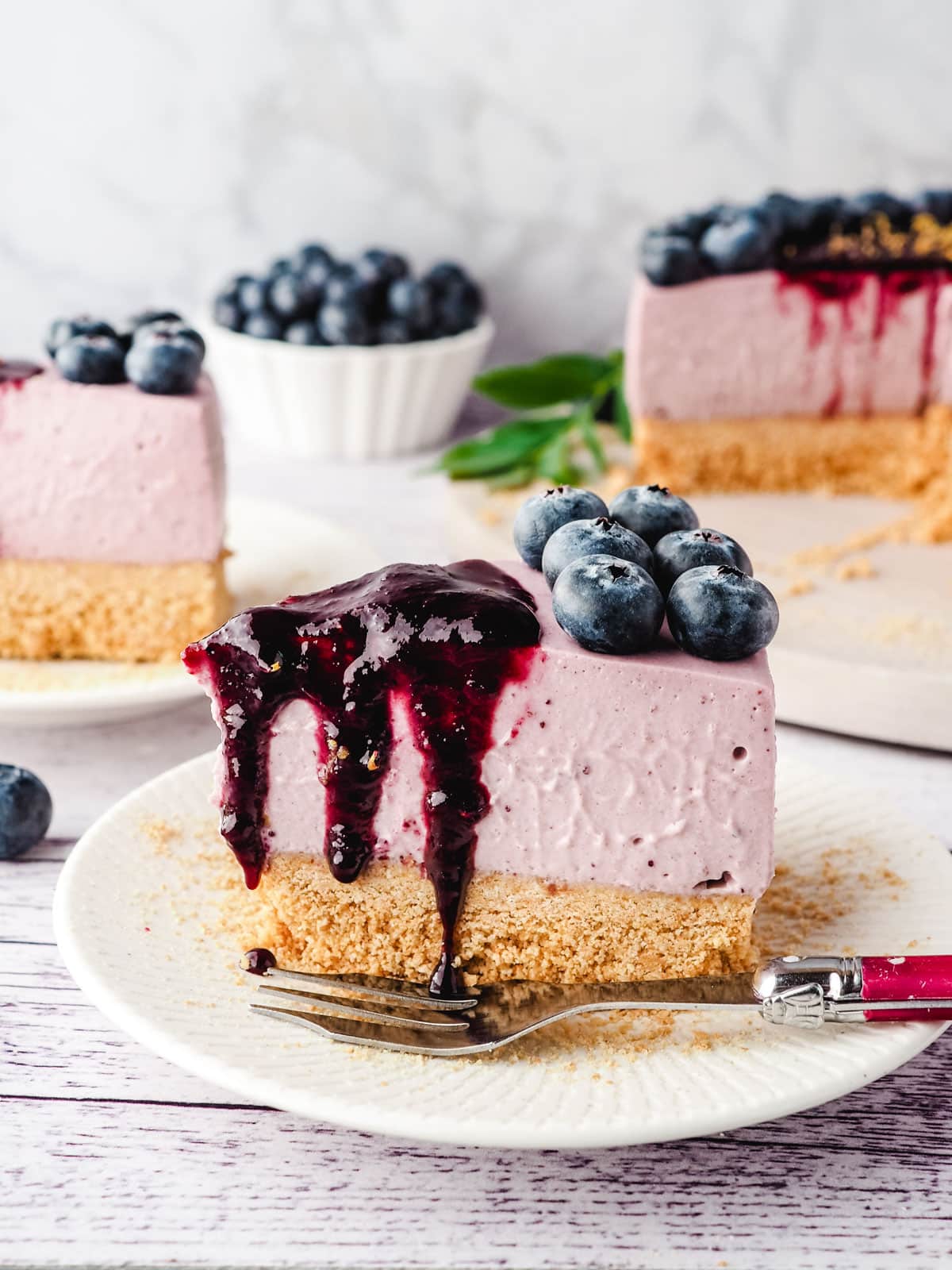 Slice of blueberry cheesecake with blueberry coulis, with another slice of cake, rest of cake and fresh blueberries in the background.