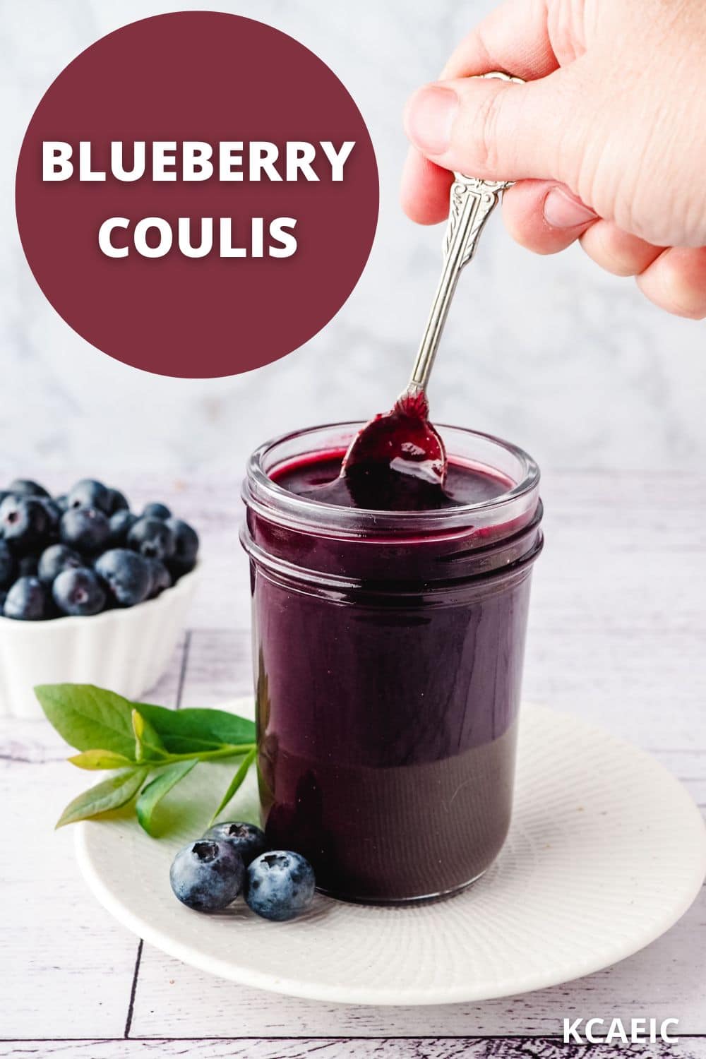 Blueberry Coulis 10 Spooning blueberry coulis from a mason jar, on a plate with fresh blueberries in the background.