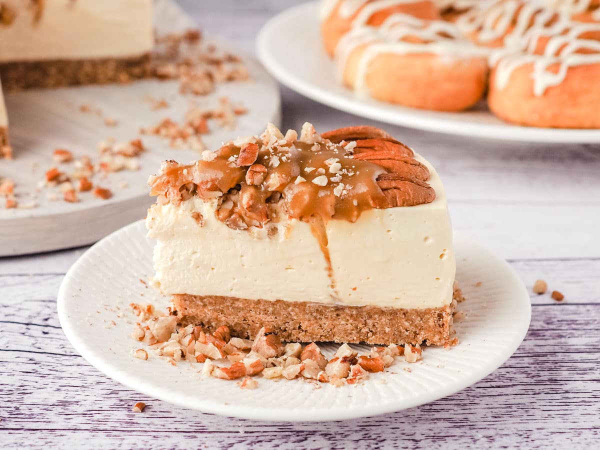 Slice of caramel pecan cheesecake on a plate, with rest of cheesecake, and a plate of air fryer cinnamon scrolls in the background.