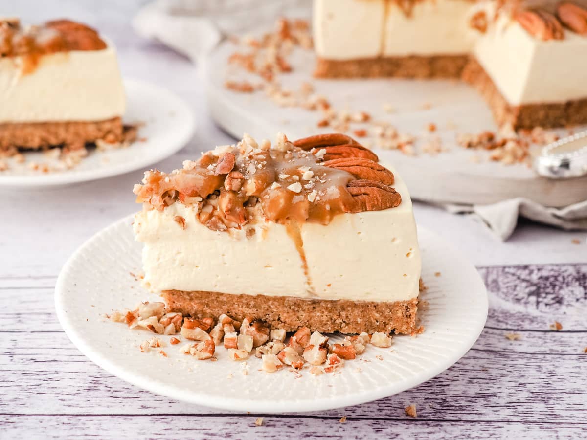 Slice of caramel pecan cheesecake on a place, with rest of cheesecake and another slice in the background.