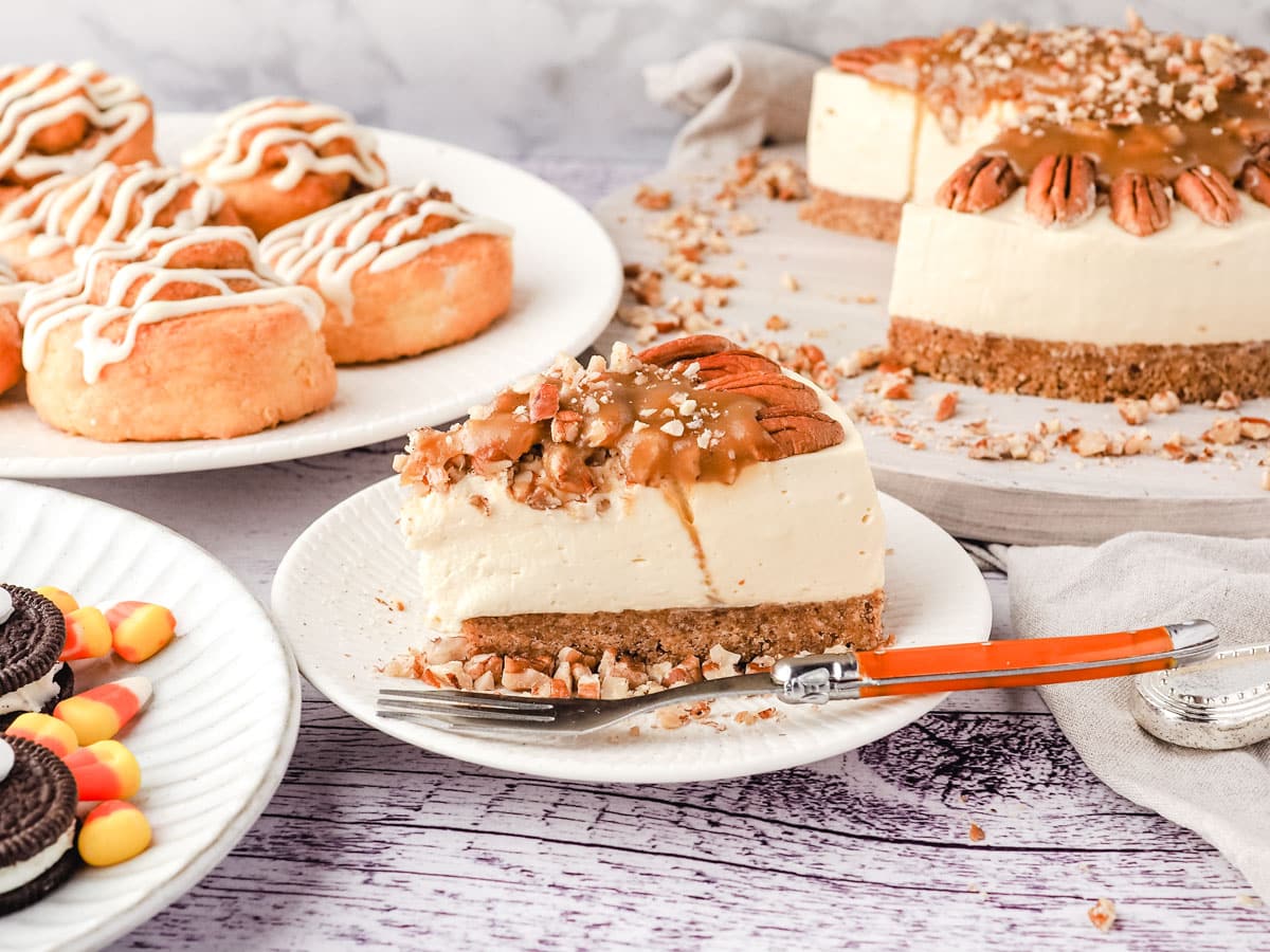 Slice of caramel pecan cheesecake on a plate, with rest of cheesecake and air fryer cinnamon scrolls and Oreo turkey cookies in the background.