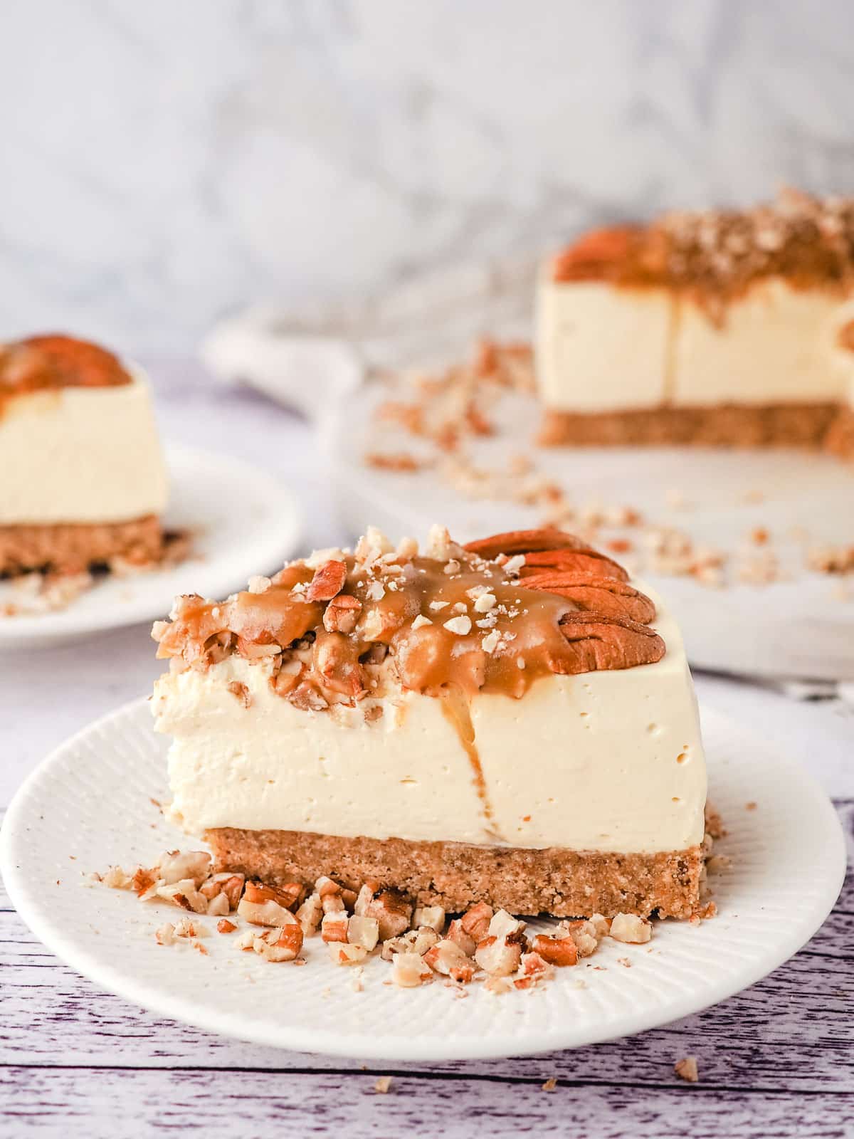 Slice of caramel pecan cheesecake on a place, with rest of cheesecake and another slice in the background.